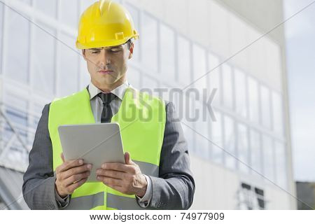 Young male engineer in reflector-vest and hardhat using digital tablet outside industry