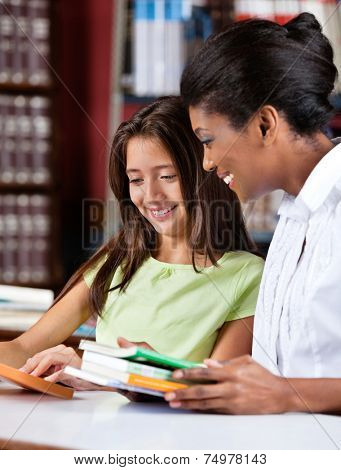 Happy female librarian and schoolgirl looking together at book in library