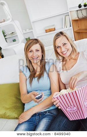 Two Attractive Female Friends Watching Televison On The Sofa Eating Popcorn