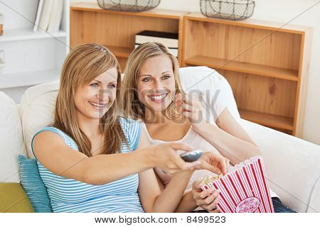 Two Bright Female Friends Watching Televison On The Sofa Eating Popcorn