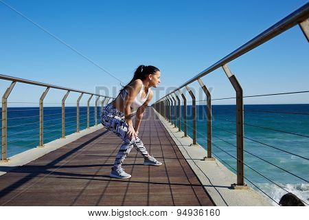 Sporty woman stretching legs while standing on wooden jetty with ocean horizon on background