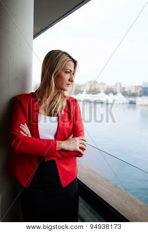 Pensive businesswoman looking out of an office balcony with beautiful seaport view on background
