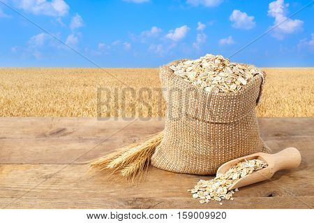 Oat flakes in sack. Ears of oats and oatmeal in bag on table with field on the background. Agriculture and harvest concept. Gold field and blue sky. Uncooked porridge