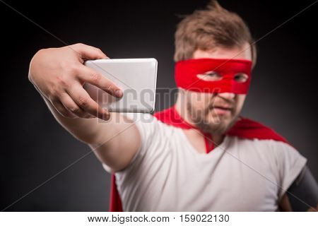 Egoist. Self. Hero. Super Hero. Picture of super hero man wth red mask on making photos on mobile or smart phone in studio.