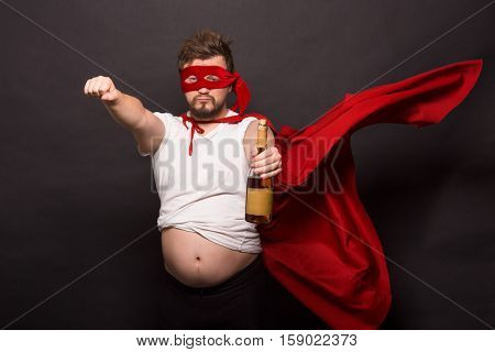 Picture of bad super hero man showing bottle with wine while posing for photographer isolated on black background in studio.