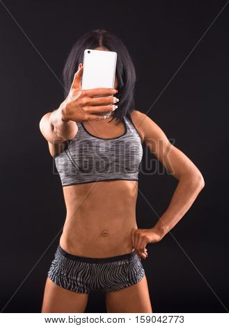 Sports, gym concepts. Brunette fitness woman making self photos on mobile or smart phone and demonstrating her fitness body isolated on black in studio.
