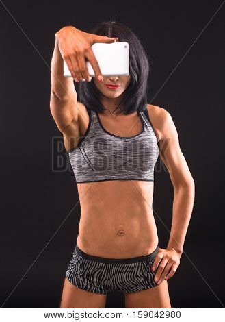 Fitness concept. Sports lady making self photos on her mobile or smart phone while posing for photographer isolated on black background in studio.