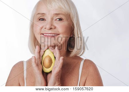 Natural cosmetics. Cheerful smiling senior woman holding an avocado fruit and looking at you while standing against the white background