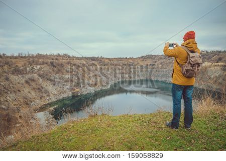 hipster guy taking photo on his mobile phone camera of lake in cold autumn day