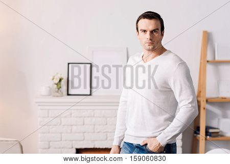 Full of confidence. Self assured handsome man holding his hand in the pocket and looking at you while standing at home