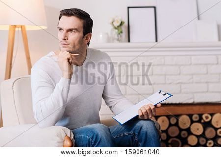 Brainstorming. Pleasant hadsome professioanl psychologist thinking and holdign folder while sitting in the armchair