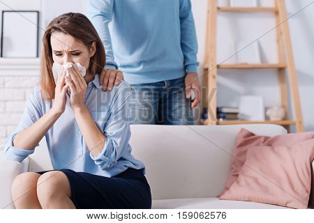 Terrible illness. Pleasant sick woman sneezing and sittign on the couch while her husband holding hand on the shoulder