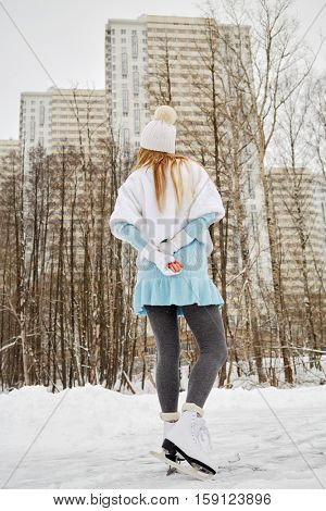 Young woman skates at outdoor skate rink in winter park against highrise buildings, rear view.