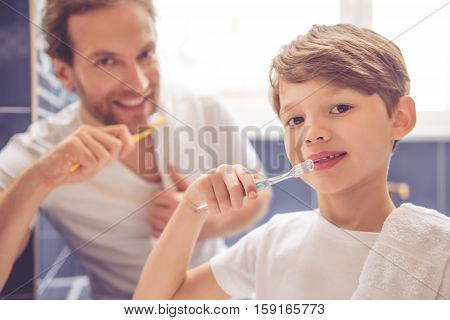 Father and son are looking at camera and smiling while brushing teeth in bathroom