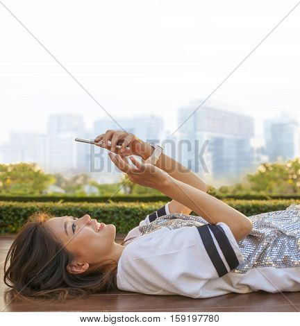 city life style of asian woman lying and touching on mobile phone screen against green environment and urban skylind background