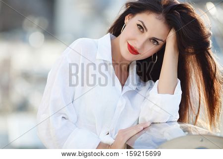 Slim beautiful brunette woman with thick long hair and brown eyes,light makeup and red lipstick in my ears,wears large gold earrings,dressed in a white shirt  and blue jeans,posing in the summer near the sea on blurred background of yachts