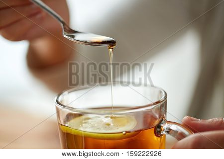 healthy food, eating and ethnoscience concept - close up of woman adding honey to tea cup with lemon