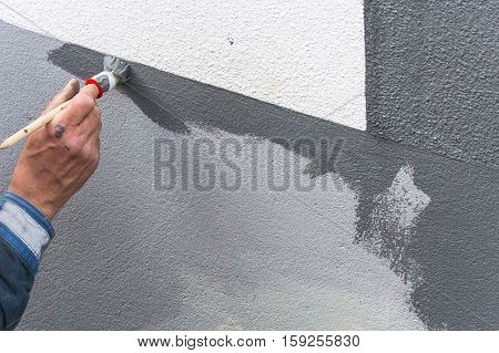 Close-up arm of a house painter with paint roller in hand painting a house wall with gray paint.