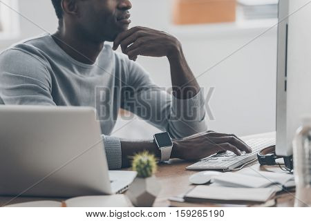 Thinking about new possibilities. Close-up of handsome African young man keeping hand to his face while sitting at his working place in home office