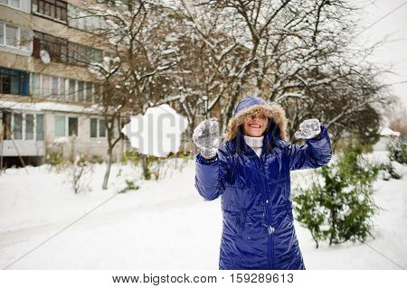 Young woman throws snowball. She aims in someone and laughs. She is dressed in a bright blue down-padded coat with a hood. It is snowing. Ground is covered with snowdrifts. Woman likes such weather.