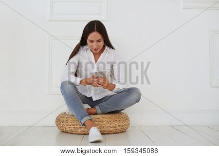 Nice pleasant concentrated woman sitting on a hassock and using cell phone while texting with colleagues