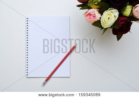 A mid shot of white jotter and a red pencil placing next to the circle of peonies. Objects against the sweet white background