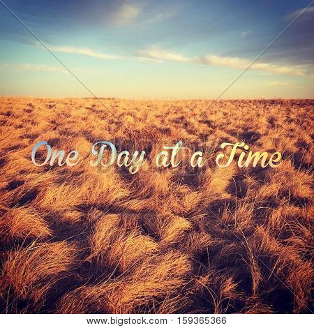 Inspirational quote.  One day at a time. Bright colorful field landscape with blue sky, clouds and orange colored grass in prairie field at sunset.    Colorful sky over horizon.  Instagram effects
