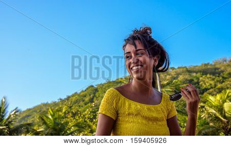 27 November, 2016. Laughing woman in yellow blouse holding her sunglasses at sunny day with blue sky and green hill on the background at Leme district, Rio de Janeiro, Brazil