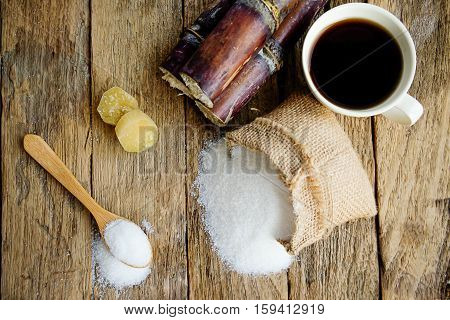 Sugar in wooden spoon with sugar in burlap sack sugarcane and white cup of coffee on wooden desk. Sugar crystals closeup. Top view and soft focus(selective focus).