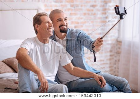 Selfie with my love. Smiling positive cheerful homosexual couple sitting in the bedroom and holding the selfie stick while expressing happiness and taking selfie