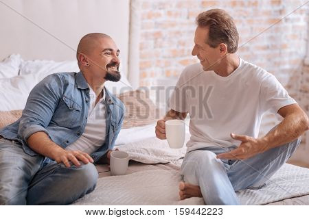 Funny time together. Smiling involved cheerful non-traditional couple sitting on the bed and drinking tea while expressing happiness and having conversation