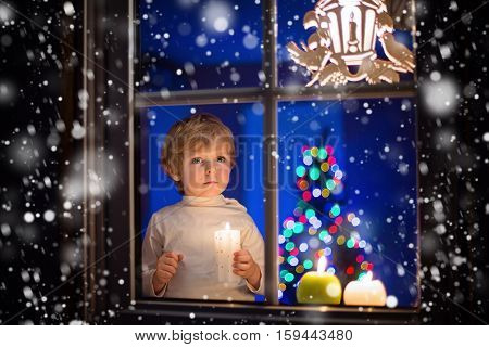 Beautiful little kid boy, standing by window at Christmas time and holding candle. Child waitng for Santa. With colorful lights from Christmas tree on background.