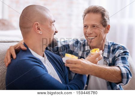 We having breakfast . Delighted smiling cheerful homosexual couple sitting on the floor in the bedroom while having fun together and feeding each other