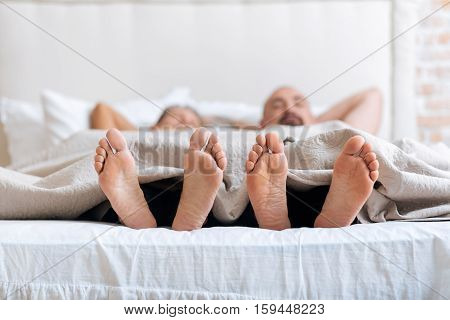 We showing our feet. Cheerful positive pleasant gay couple lying on the bed in the bedroom while expressing love and care and showing their feet under the blanket