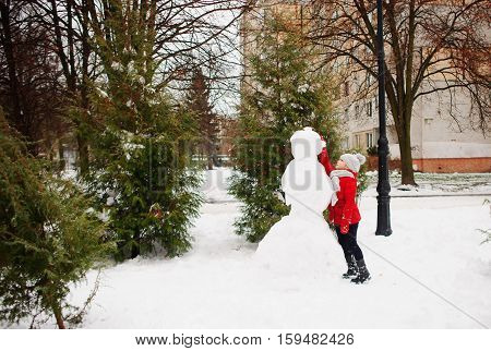 The girl of school age builds a snowman. On the ground there is a lot of snow. The girl in a red jacket and a gray knitted cap stands near a big snowman. She decorates the head of a snowman.