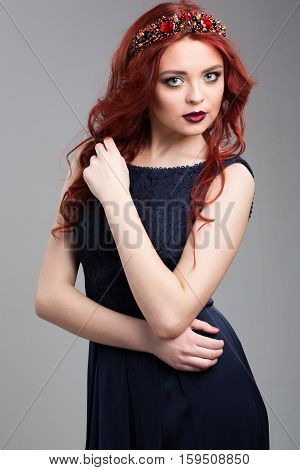 Beautiful red-haired fashion model posing in evening dress and in the diadem over dark background. female gestures of seduction. body language. touching her hair.