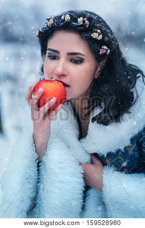 Portrait of young woman dressed in blue coat. She is walking in the winter forest holding a basket with apples