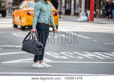 Elegant man walking on city street crosswalk wearing casual clothes with jeans ant t-shite and holding travel bag and sunglasses in hands