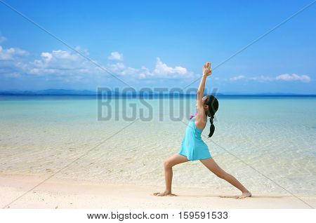 Young healthy Asian kid doing yoga on tropical beach in warrior pose I. Preteen girl in swimwear practice yoga on sea beach blue sky sand sun daylight relaxation landscape for sun protection ad.