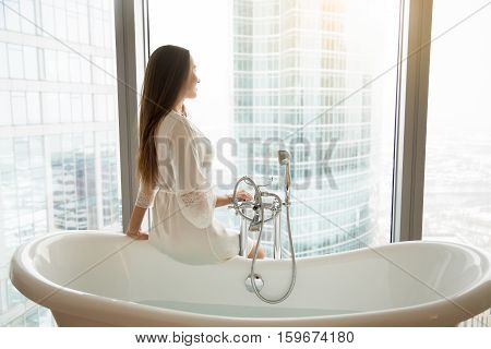Profile portrait of young model in a gown sitting at the side of freestanding white bath, bathroom design, luxury sanitary ware collection, preparing relaxing bath. Beauty, healthy lifestyle concept