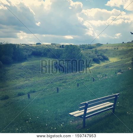 Empty wooden isolated park bench overlooking lush green hills of trees and grass.Wooden fence posts in front of bench.Blue sky with big white and grey clouds in background.