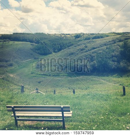 Empty wooden isolated park bench overlooking lush green hills of trees and grass with trails through valley.Wooden fence posts in front of bench. White and grey clouds in background. Lighting effects.