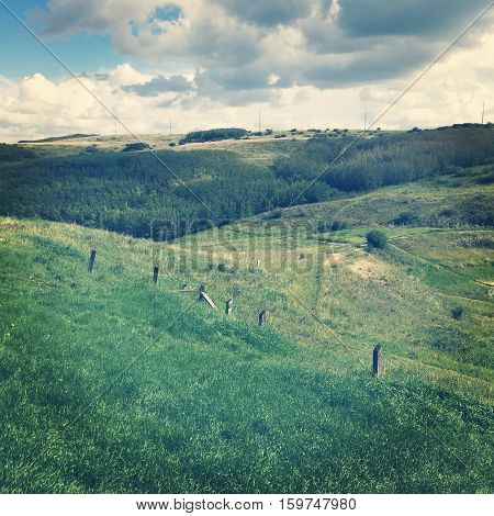 Field landscape in summer.  Lush dark green valley and hills with old wire fence and posts, bright lush green trees and grass. Bright blue sky, dark and white clouds background.