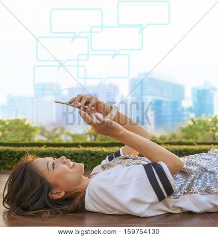 city life style of asian woman lying and touching on mobile phone screen against green environment and urban skylind background