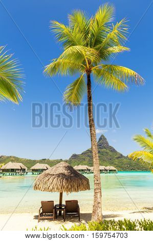 Two sunlounger chairs under a thatched parasol on a sand beach with a view on the lagoon and Mount Otemanu on the tropical island of Bora Bora near Tahiti in French Polynesia.
