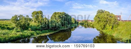 Summer warm evening. Panorama with river views from the footbridge. The village is in the background. Reflection of blue sky in the river water.