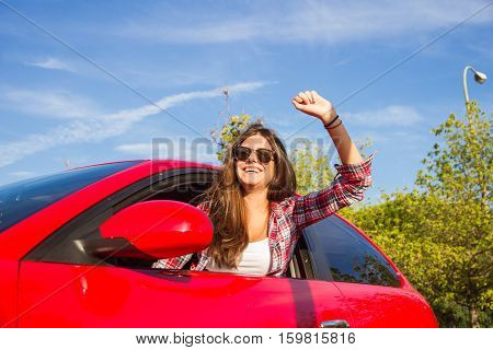 Portrait of happy young woman going on a road trip leaning out of window of the red car. She has sunglasses on. She is happy.