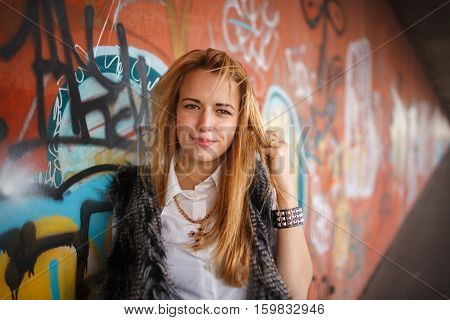 Russian beautiful smiling teenager girl with long blond hair and make up near the wall with graffiti, selective focus