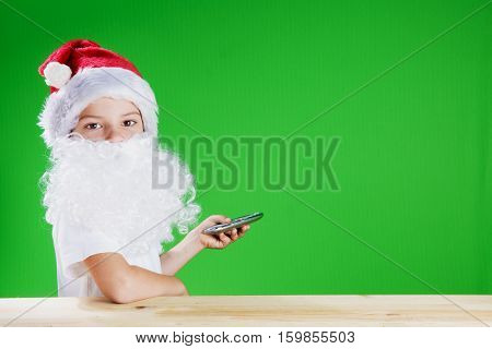 Little Santa Claus looking into the camera. He presses the button on the remote control. Remote is aimed at green background. Сhromakey. Close-up.