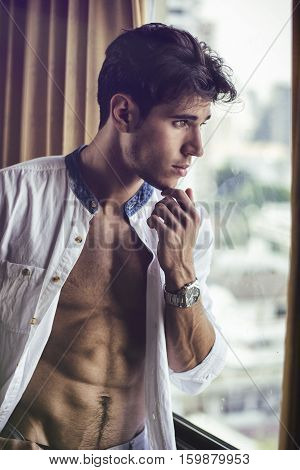 Sexy young man standing with shirt open on muscular chest in his bedroom next to window curtains